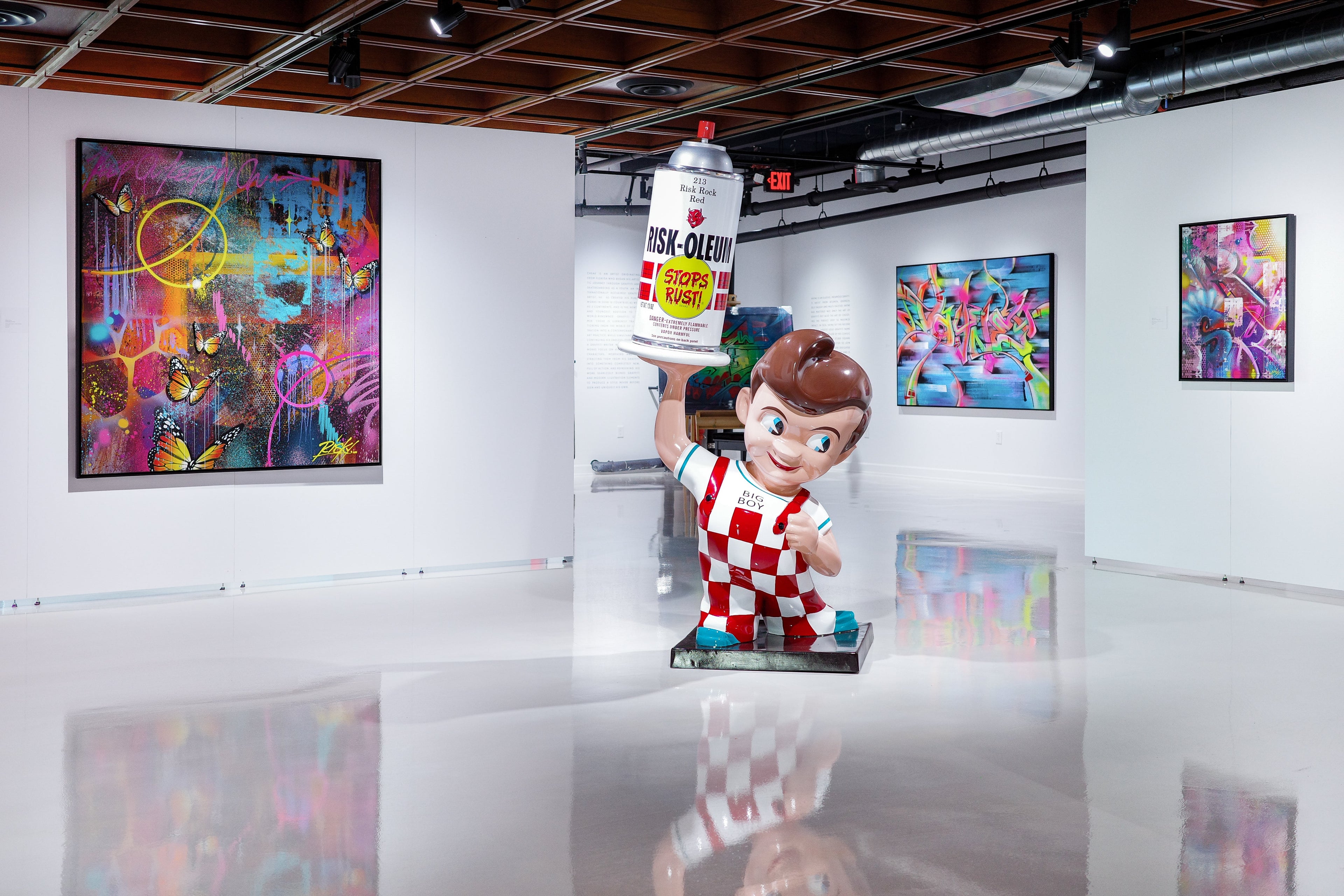 Birds of a Feather sculpture: boy in red-and-white checkered outfit lifts an oversized spray can in a bright gallery.